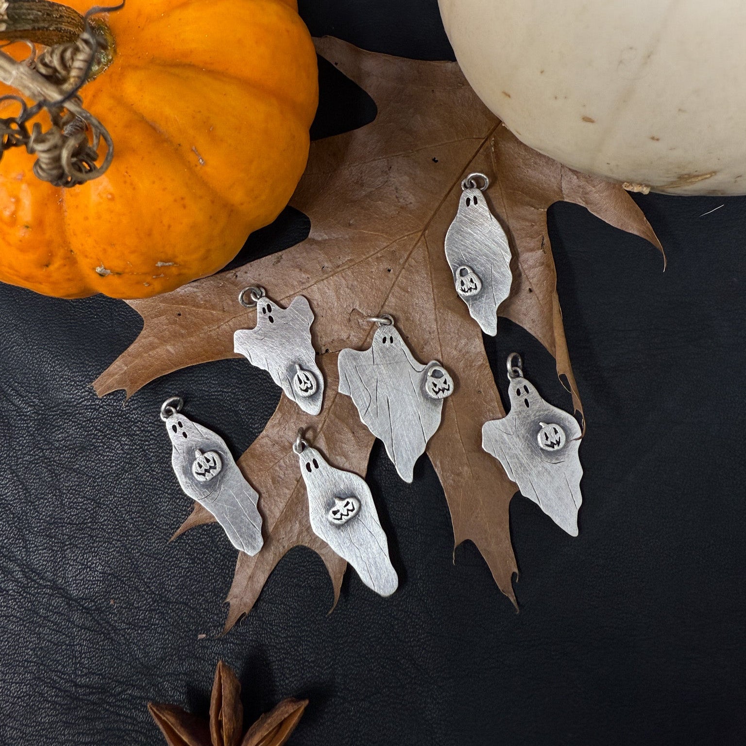 Silver ghost-shaped pendants on a dark surface with pumpkins and leaves